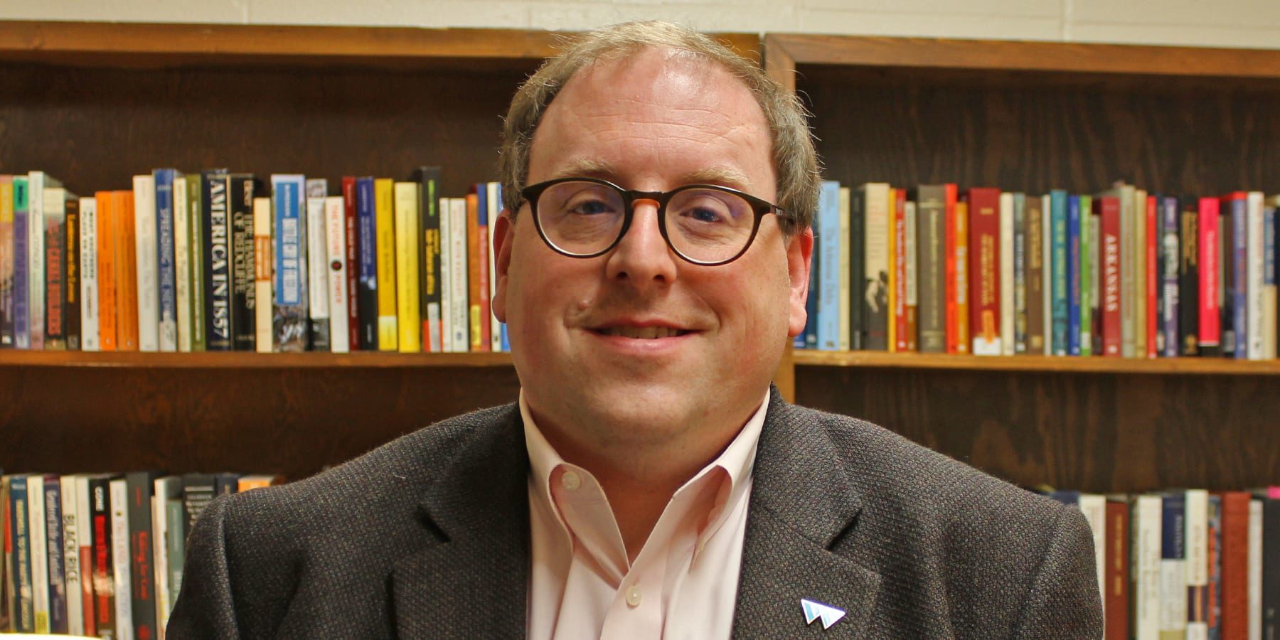Photograph of a man wearing glasses and a sport coat in a library.
